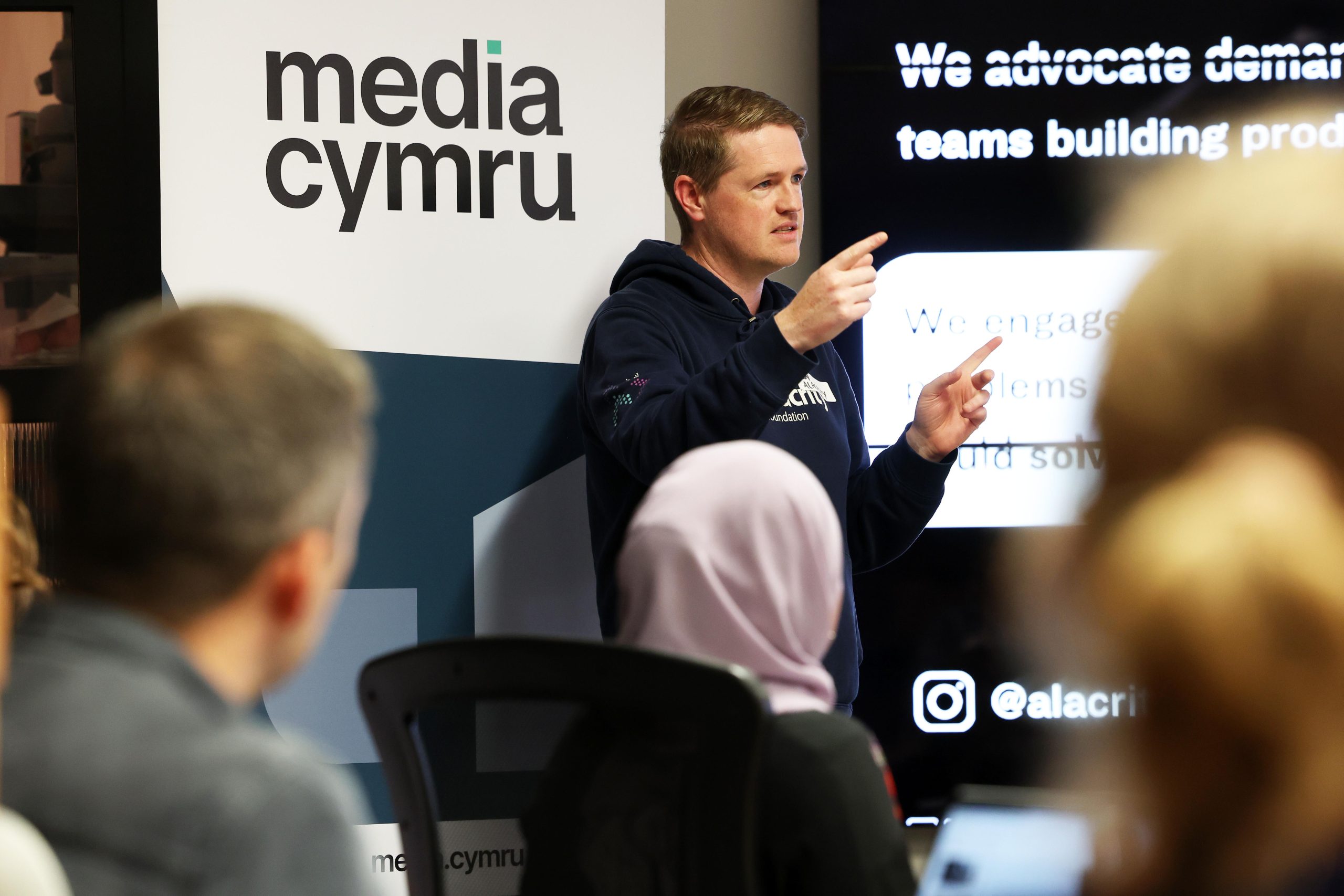 man talking to an audience a media cymru logo is behind him