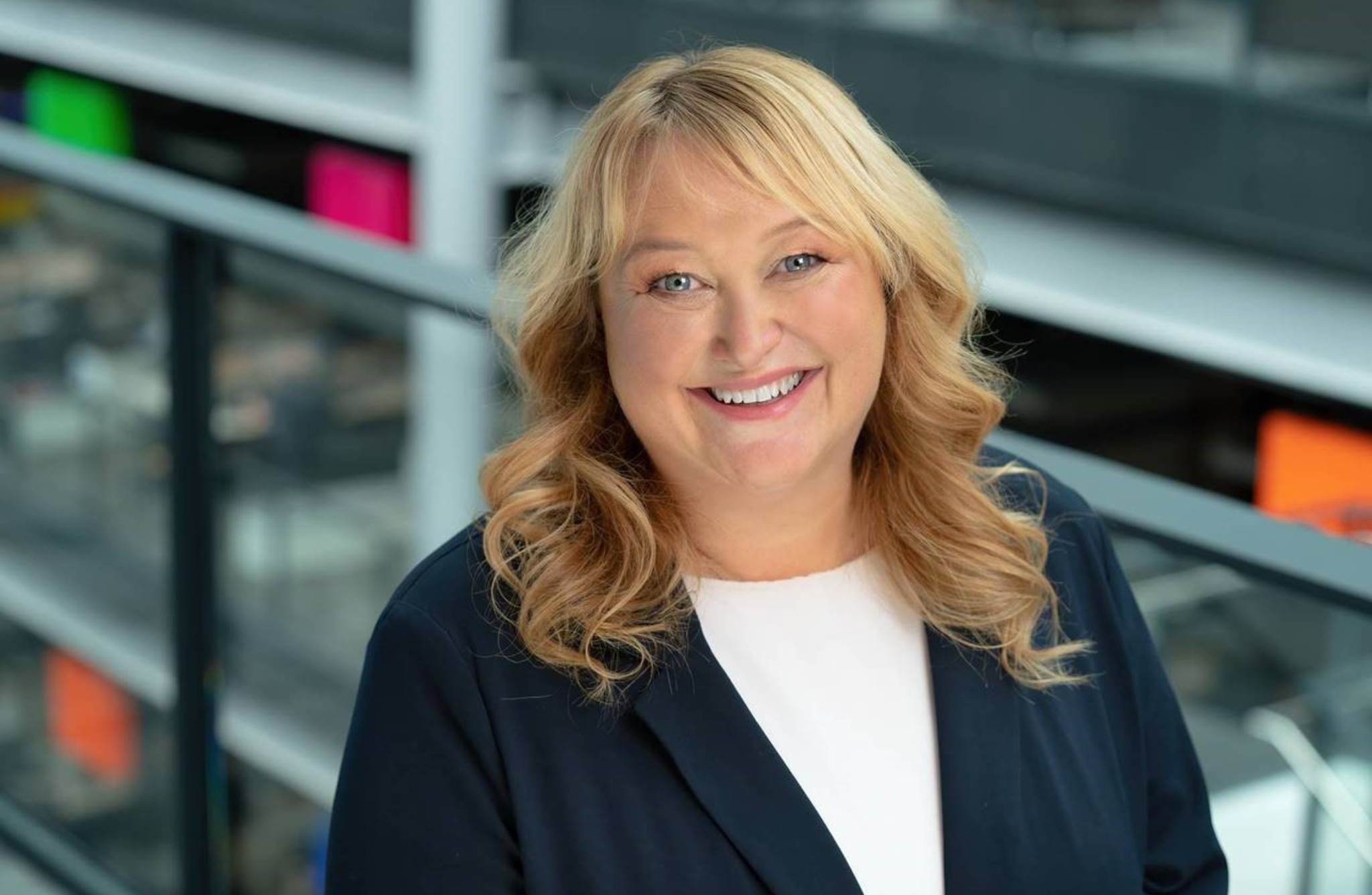 Mae person â gwallt melyn hir, tonnog yn sefyll o flaen rheiliau gwydr. Mae'r cefndir yn dangos lleoliad dan do gyda phaneli lliwgar amrywiol a dyluniad pensaernïol modern. Mae'r person yn gwisgo siaced dywyll dros dop gwyn.