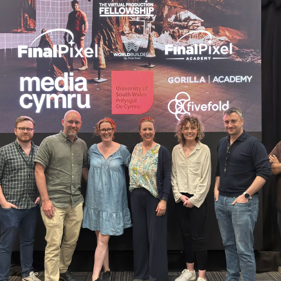 A group of eight people standing in front of a large screen displaying various logos and text. The screen shows the following: 'Final Pixel,' 'media cymru,' 'University of South Wales Prifysgol De Cymru,' 'THE VIRTUAL PRODUCTION FELLOWSHIP WORLDBUILDERS,' 'Final Pixel Academy,' 'GORILLA | ACADEMY,' and 'fivefold.' The individuals are dressed in casual attire.