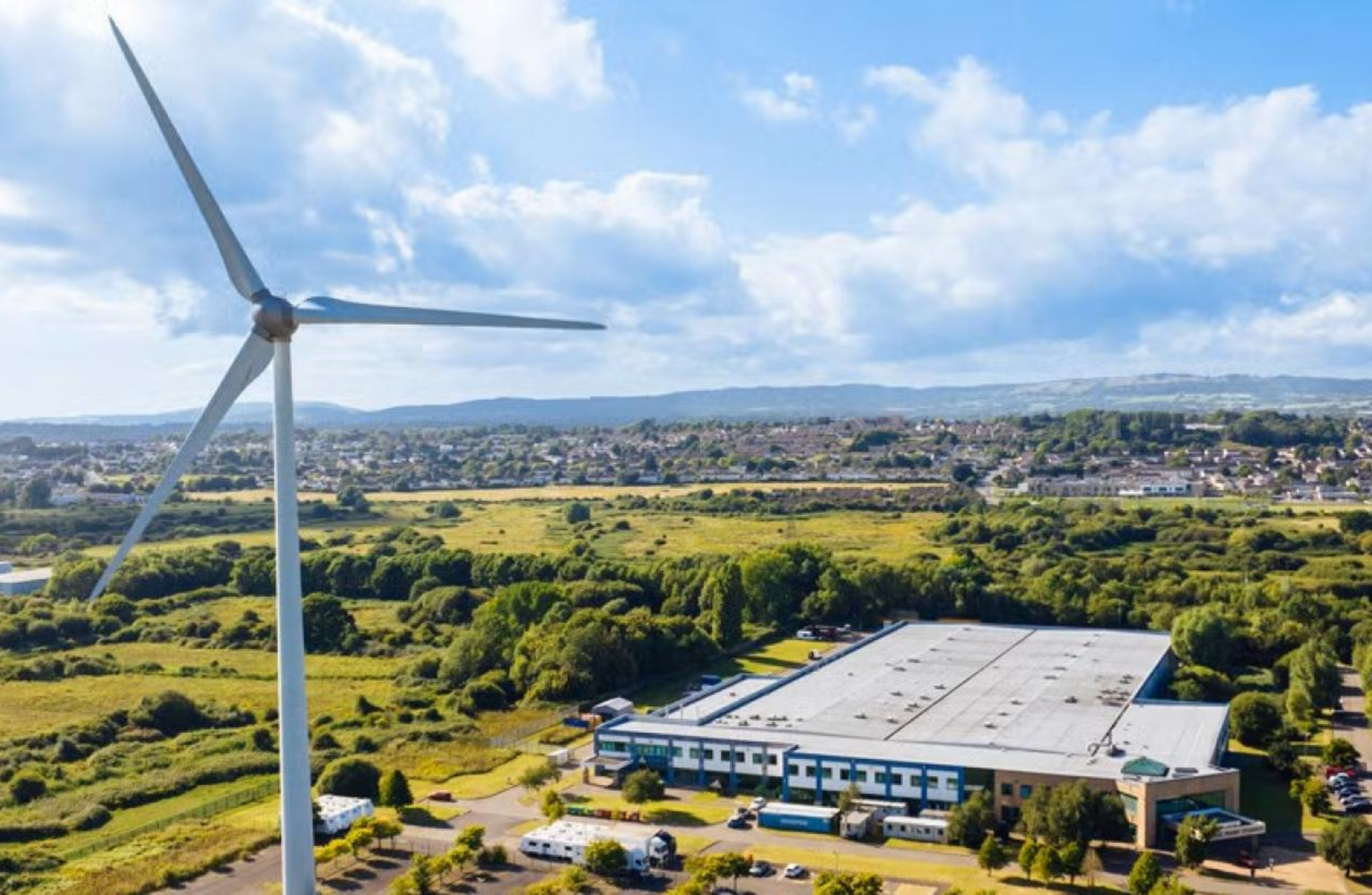 A large wind turbine with three blades stands prominently in a green landscape. Behind it, an industrial building with a flat roof is surrounded by trees and fields. Further back, a residential area with houses is visible, with hills in the distance under a partly cloudy sky.