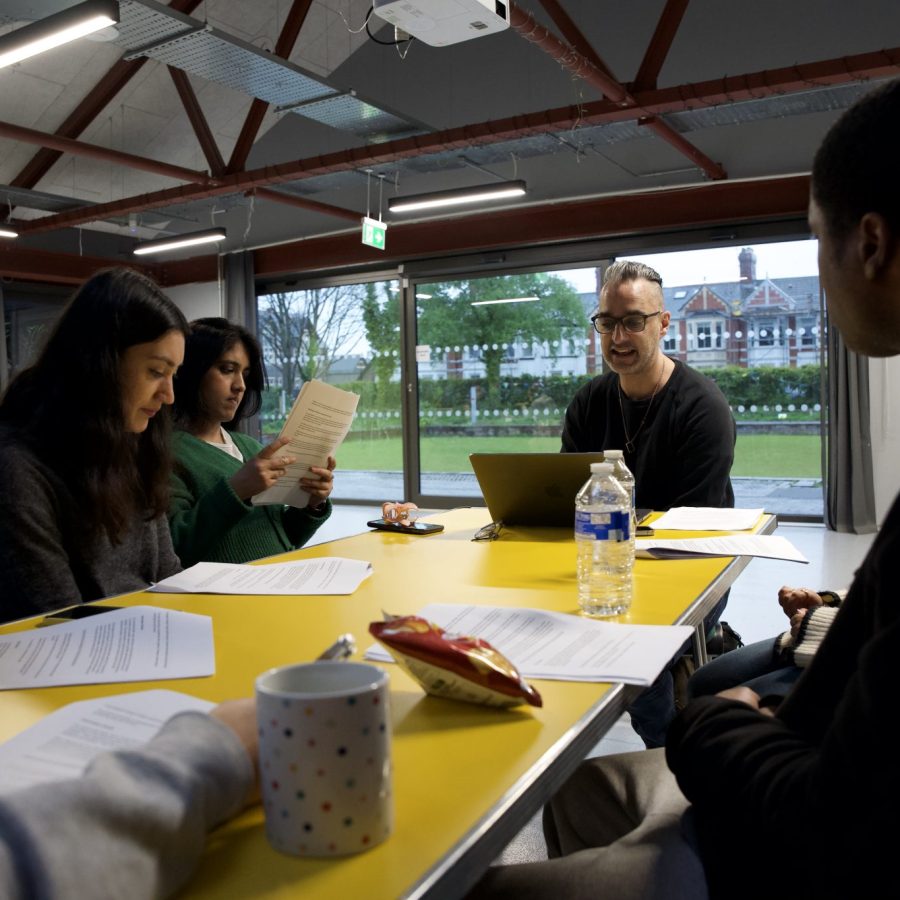 A group of people seated around a yellow table in a modern indoor space with large windows overlooking a green lawn and a building in the background. Papers, a laptop, a water bottle, a mug, and a snack packet are on the table. The ceiling has exposed beams and lighting fixtures, and a projector is mounted above.