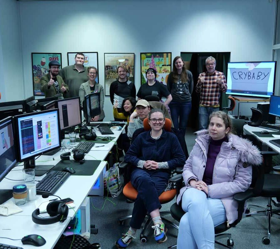 A group of people gathered in an office space filled with multiple computer workstations. Several monitors display colorful graphics and software interfaces. The room has bright lighting, white walls, and framed posters on the back wall. Two individuals are seated in orange chairs at the front, while others stand behind them near the desks. Various office items, such as keyboards, headsets, and a coffee cup, are visible on the desks.