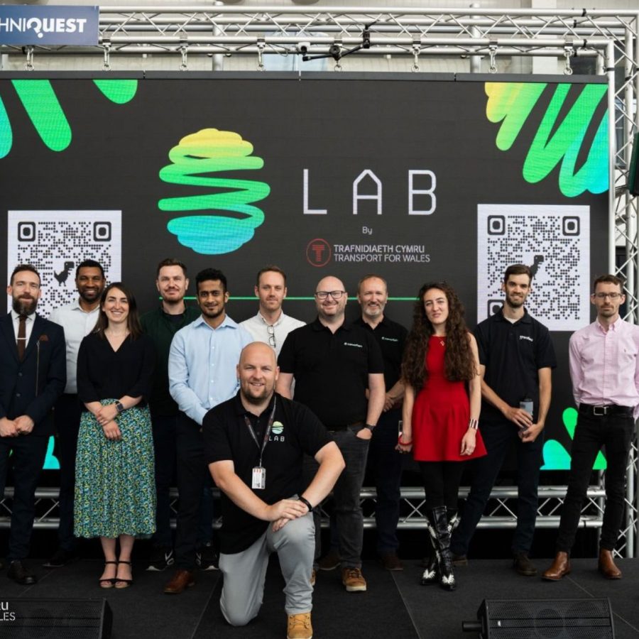 A group of people standing and kneeling on a stage in front of a large digital screen displaying the word “LAB” with a stylized green and yellow abstract logo. The screen also shows two QR codes and the text “Transport for Wales” in English and Welsh.