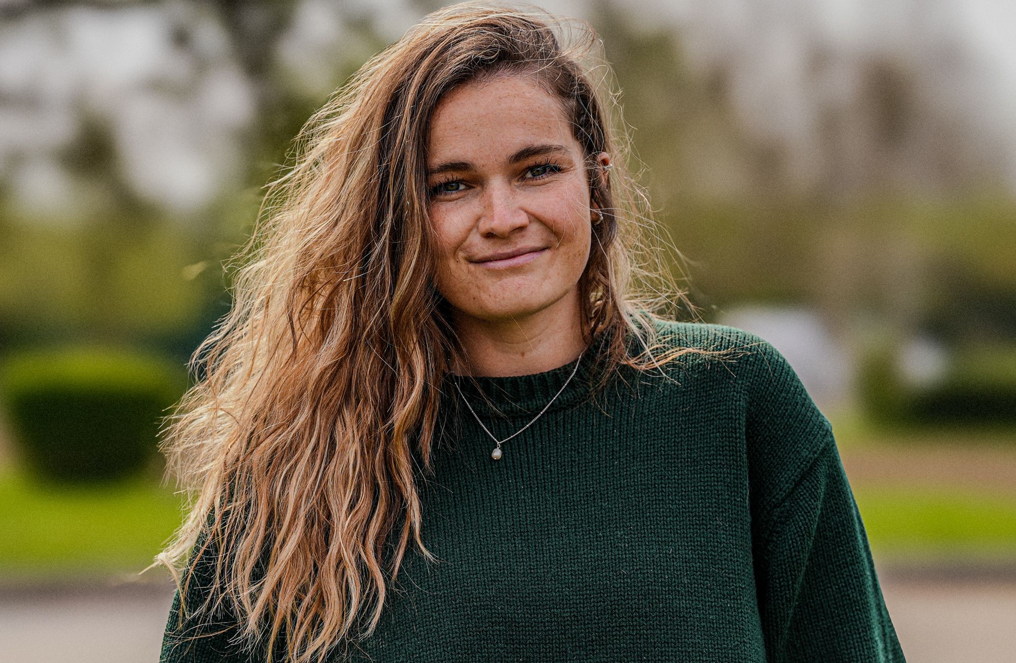 A person wearing a dark green knitted sweater with a simple necklace featuring a small pendant. The individual has long, wavy, light brown hair cascading over the shoulders. The background is outdoors with blurred greenery and soft lighting.