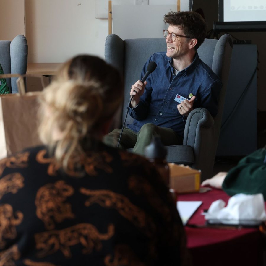 Four people are seated indoors around a red table during a discussion. One person in a green outfit faces another in a gray armchair holding a microphone. Two others are partially visible in the foreground. A paper bag, notebooks, and a projection screen are also present.