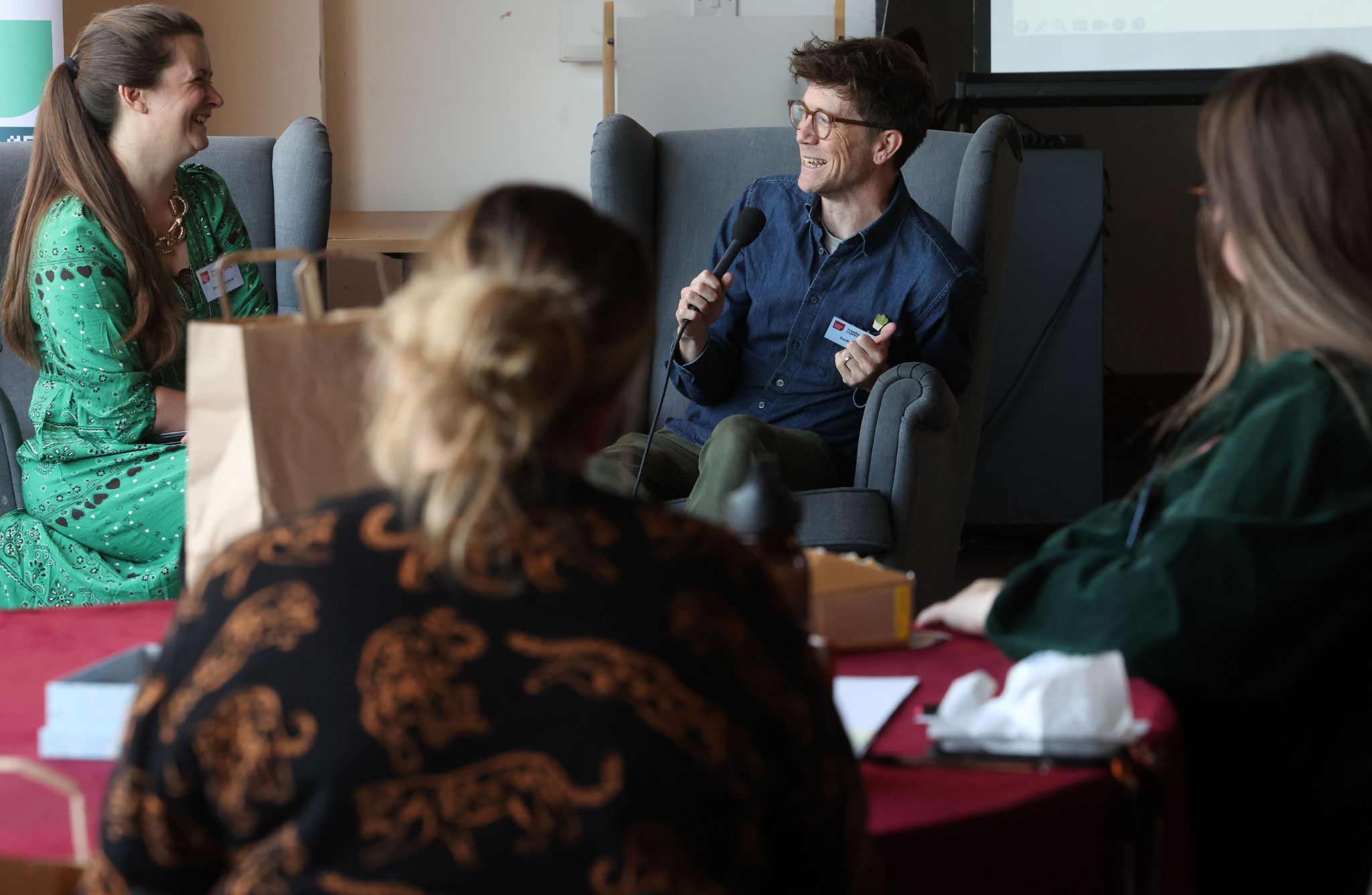 Four people are seated indoors around a red table during a discussion. One person in a green outfit faces another in a gray armchair holding a microphone. Two others are partially visible in the foreground. A paper bag, notebooks, and a projection screen are also present.