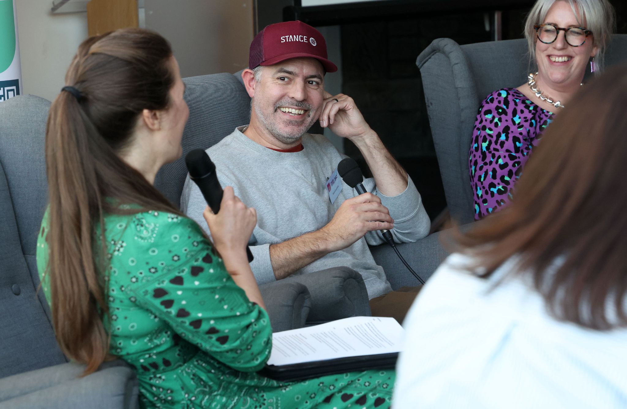 Three people are seated indoors during a discussion. One person in a green patterned outfit holds a microphone and a document, speaking to another wearing a maroon cap and gray top who also holds a microphone. A third person in a purple patterned outfit is partially visible.