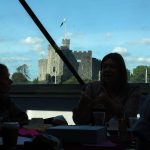 People sit at a table indoors with drinks and papers, while Cardiff Castle is visible outside through large windows under a bright blue sky.