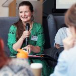 Person in a green patterned outfit speaks into a microphone while seated, holding papers. Others sit nearby with drinks on the table in the foreground.
