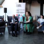Four people sit in gray chairs during a panel discussion. A banner reading “Media Cymru” and a projection screen are visible in the background.