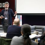 Two people stand at the front of a room presenting to an audience. One holds a phone while speaking. Attendees sit at tables facing a screen and BBC Sounds-branded wall.