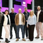 A group of nine people standing together in an open indoor space with large screens in the background displaying “BBC iPlayer.”