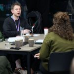 A small group sitting at a table engaged in discussion. Notebooks, drinks, and lanyards are on the table, and coiled cables are visible in the background.