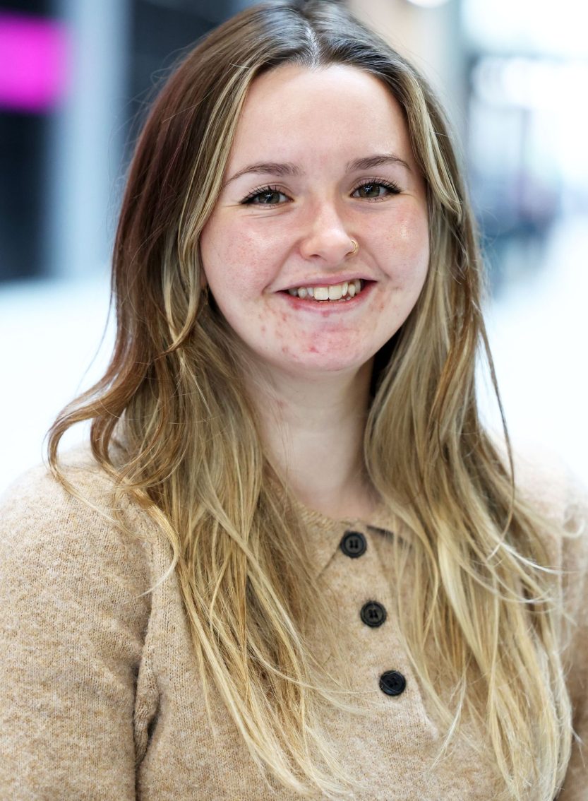 A person with long, wavy, light brown and blonde hair wearing a tan knit sweater with three dark buttons. They are standing indoors in a softly lit space with a blurred background.