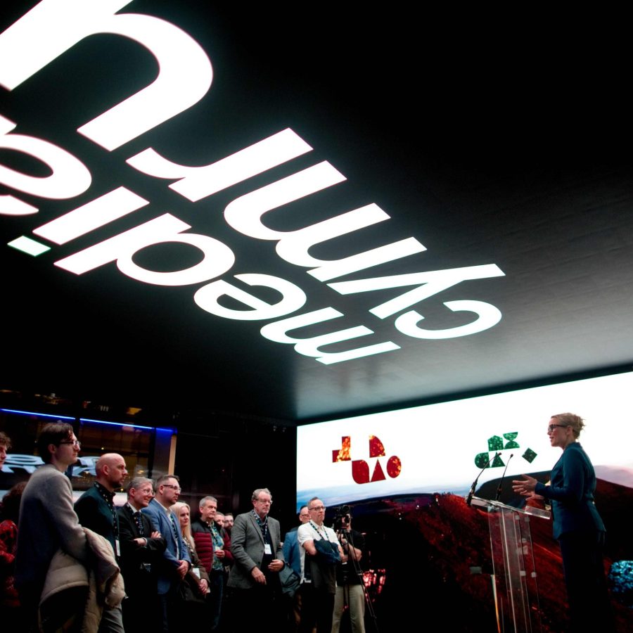 A person stands at a clear podium on a stage, speaking to an audience gathered in a dimly lit event space. A large illuminated screen behind the speaker displays abstract colorful shapes over a landscape image. Above the crowd, a ceiling installation features large, inverted white text reading “Media Cymru.” Members of the audience stand in a semi-circle facing the stage.