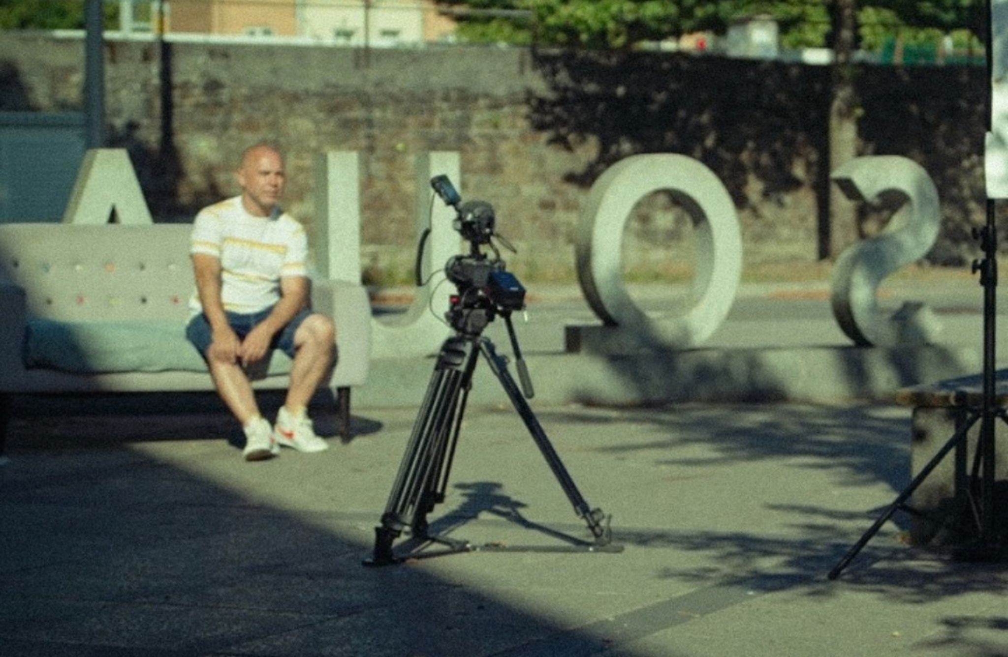 A person sits on an outdoor sofa in a shaded area while a professional video camera on a tripod is positioned in front of them. Large sculptural letters are arranged behind the scene, and trees and a stone wall are visible in the background.