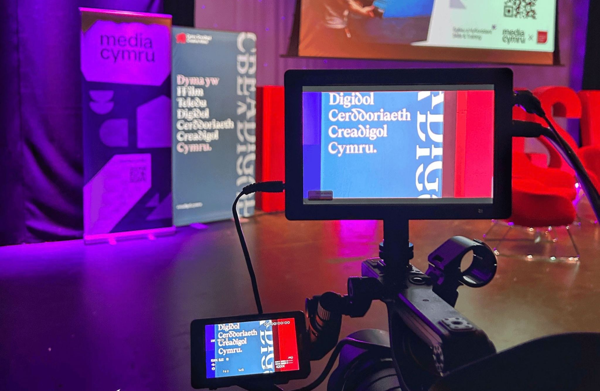 “A video camera records a stage setup with Media Cymru banners and a screen displaying Welsh text at an event.”