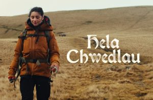 Person walking across open countryside wearing outdoor gear, with the text “Hela Chwedlau”.