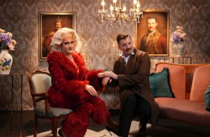 Two people seated in a vintage-style living room with framed portraits and a chandelier.