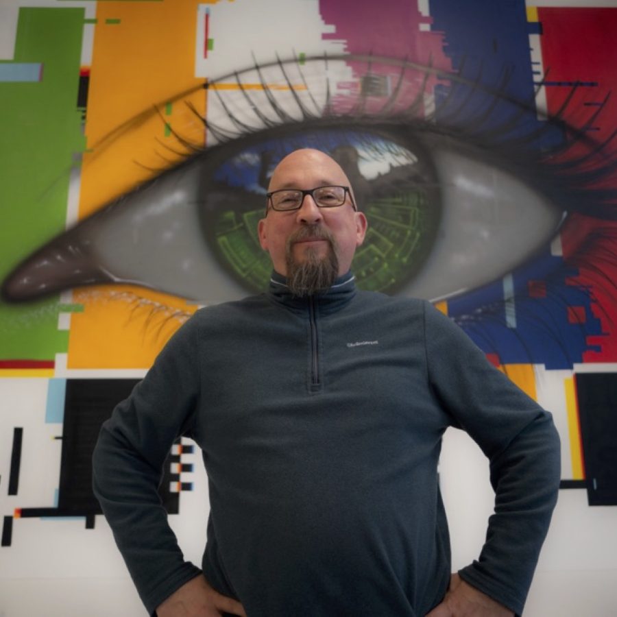 Photo of Alex Counsell, a bearded man in glasses, standing confidently in front of wall art of a giant eye.