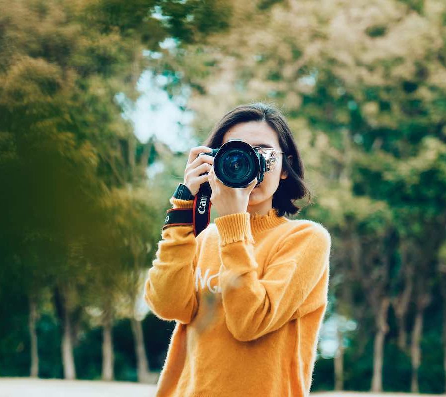 A female photographer holding up a camera while wearing an orange sweater and standing in front of a green leafy woodland background.