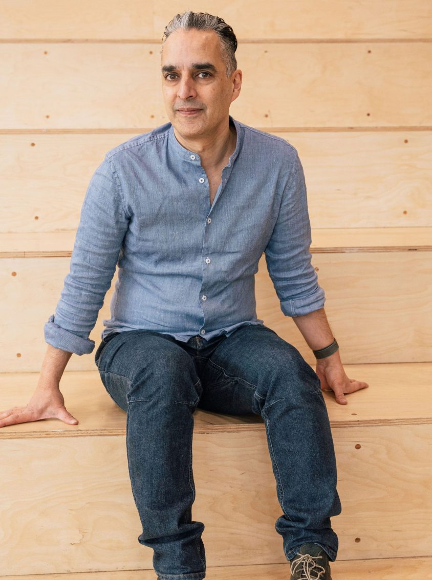 Clean shaven man with grey-blue shirt looking intent while sitting on wooden stairs.