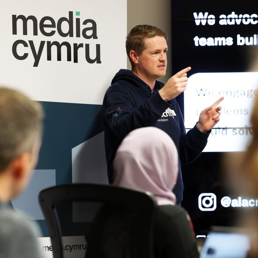 man talking to an audience a media cymru logo is behind him