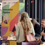 people chatting to each other, a banner with title in black saying 'Innovation Pipeline' on it. The banner has yellow, green, red and purple shapes on it.