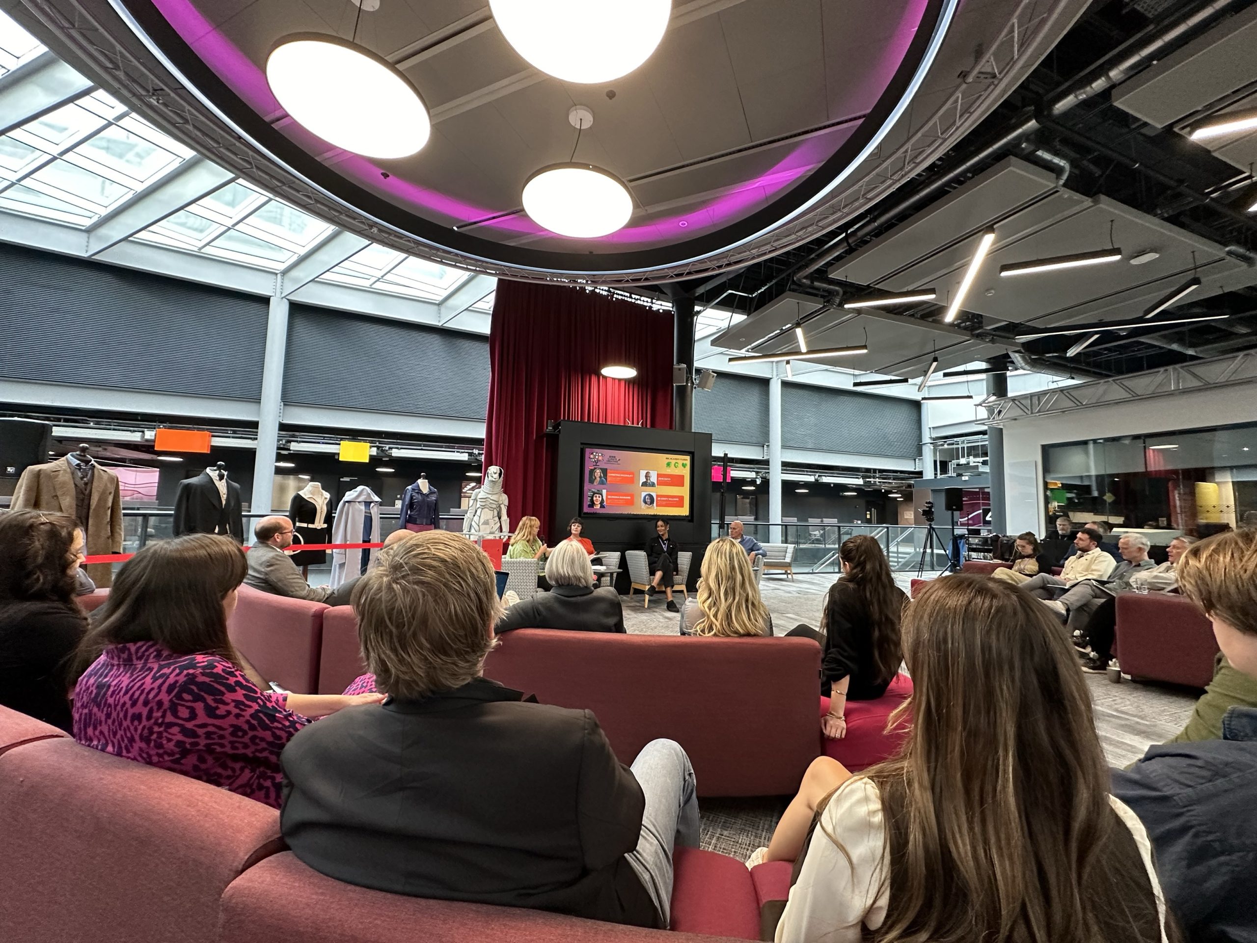 audiences of people sitting on red sofas watching four people sitting on chairs behind a TV screen