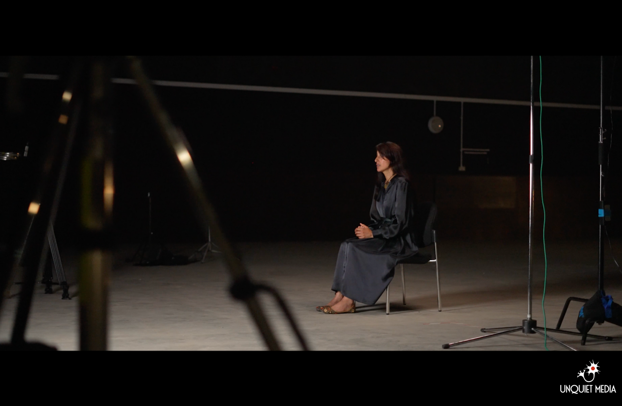 One person is sitting on a black chair in a dimly lit space. There is filming equipment around them. Logo of "UNQUIET MEDIA" is visible in the bottom right corner.