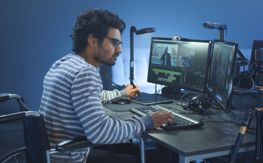 a person in a wheelchair working at a desk at his computer, the wall in the background is blue, the computer and desk is black