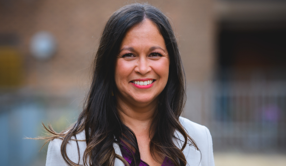 Profile of a woman's face, she is looking at the camera smiling, she has long dark brown hair and is wearing a light grey blazer