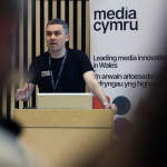 a man with dark short hair is wearing a black t-shirt and standing behind a wooden podium presenting to an audience, behind him there is a white banner with black text which reads media cymru