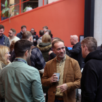 a group of people standing in an event space and networking and chatting to each other, many are smiling there is a red wall in the background