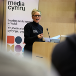 a woman with short blond hair standing behind a wooden podium presenting to an audience, she is wearing black glasses and a black t-shirt behind her there is a white banner with black text that reads media cymru