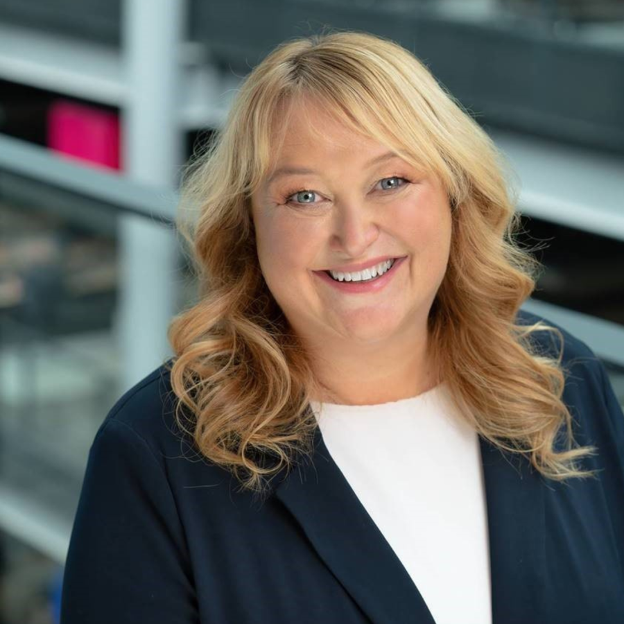 A person with long, wavy blonde hair is standing in front of a glass railing. The background shows an indoor setting with various colored panels and a modern architectural design. The person is wearing a dark blazer over a white top.