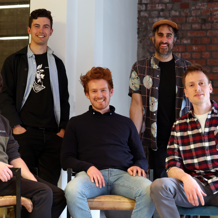five people posing the photo, smiling into camera. Three are seated at the front and two are standing at the back.