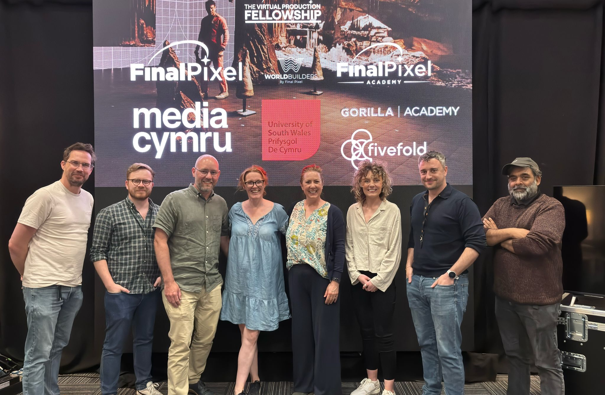 A group of eight people standing in front of a large screen displaying various logos and text. The screen shows: 'Final Pixel,' 'media cymru,' 'University of South Wales Prifysgol De Cymru,' 'THE VIRTUAL PRODUCTION FELLOWSHIP WORLDBUILDERS,' 'Final Pixel Academy,' 'GORILLA | ACADEMY,' and 'fivefold.' The individuals are dressed in casual attire