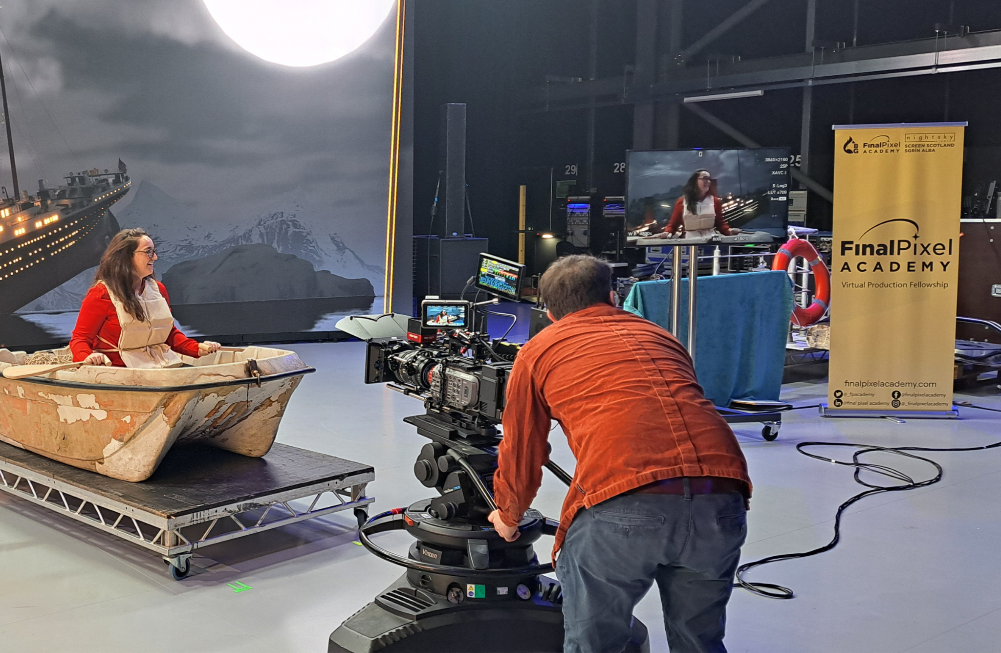Final Pixel Academy A behind-the-scenes image of a film set. A woman in a red top and white apron sits in an old, weathered boat placed on a platform. In front of her, a cameraman in an orange shirt operates a large camera mounted on a dolly. The background features a large screen displaying an image of the Titanic ship and icy mountains under a full moon. To the right, there is another monitor showing the same scene being filmed, and next to it is a yellow banner with 'FinalPixel Academy' written on it along with some contact information. Various equipment and cables are visible around the set.