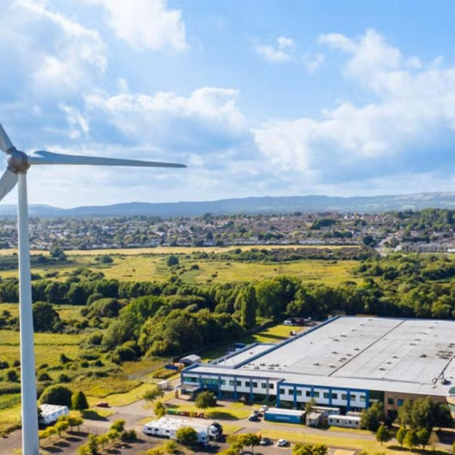 A large wind turbine with three blades stands prominently in a green landscape. Behind it, an industrial building with a flat roof is surrounded by trees and fields. Further back, a residential area with houses is visible, with hills in the distance under a partly cloudy sky.