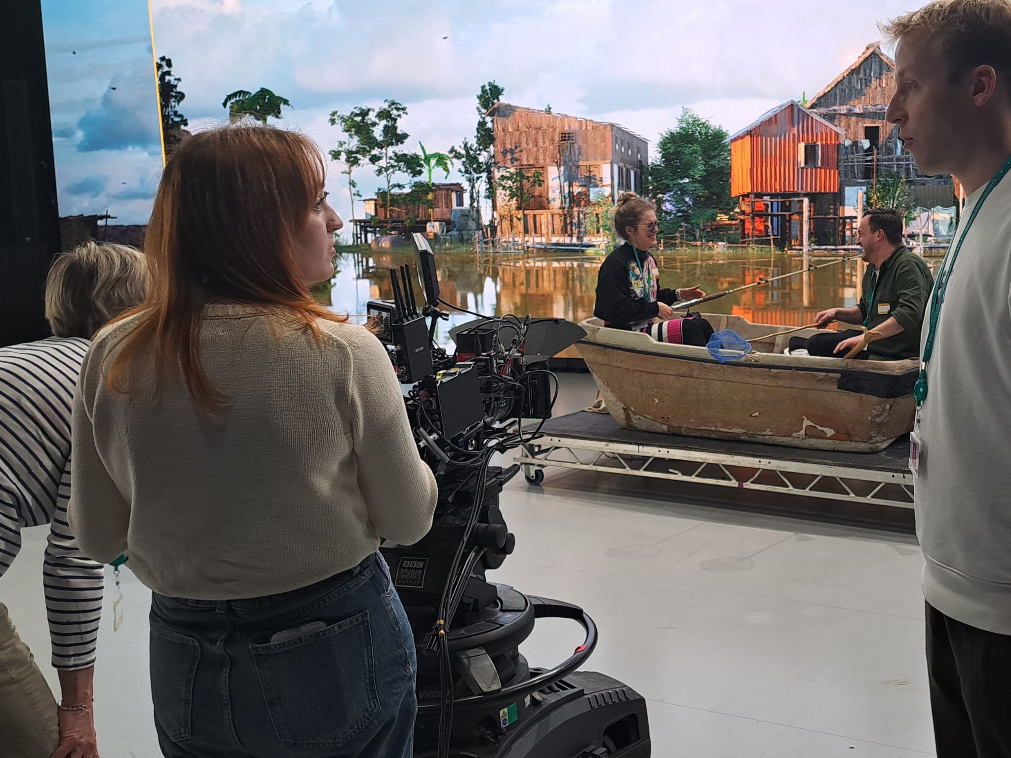 picture from a film set. 3 people stand behind a camera looking at the two people sat in a boat in front of a virtual production large screen