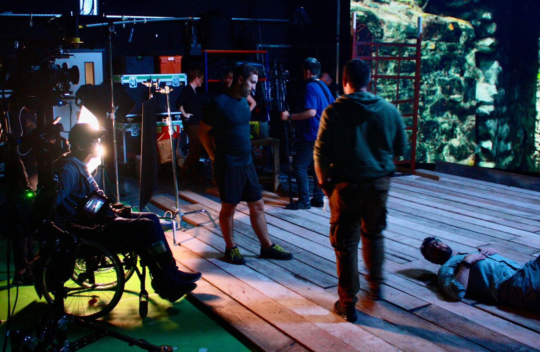 Behind-the-scenes view of a film or video production set. Several people are present, including a camera operator, a person in a wheelchair handling equipment, and others standing or sitting nearby. One individual lies on a wooden floor, possibly an actor. The background features a green screen with projected imagery resembling stone walls, surrounded by various production equipment.