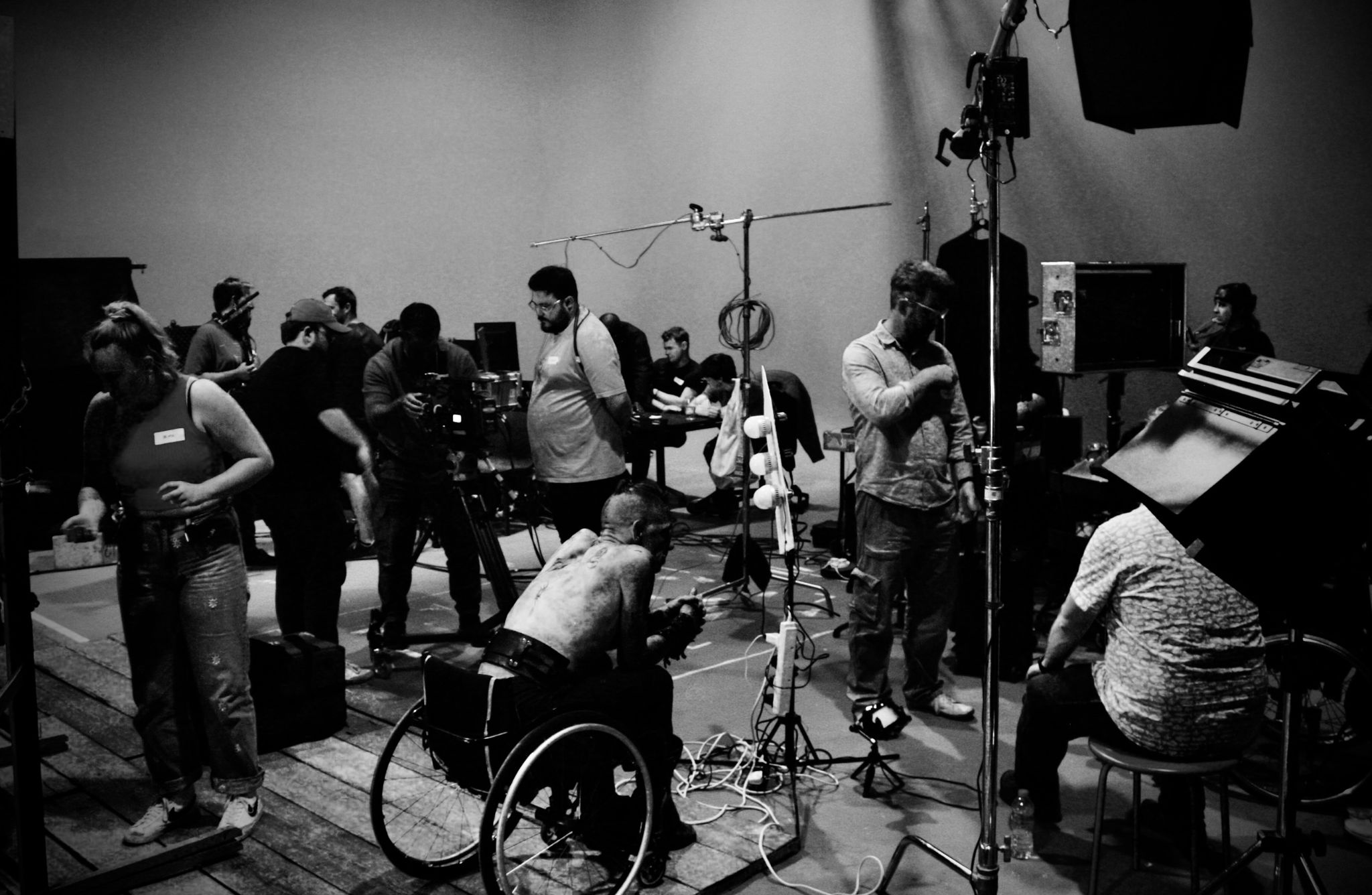 Black and white photo of a busy film or photography set. Multiple people are engaged in various tasks, surrounded by equipment including cameras, lighting rigs, and cables. One person in a wheelchair adjusts equipment, while another holds a camera. The scene captures the collaborative, behind-the-scenes effort of media production.