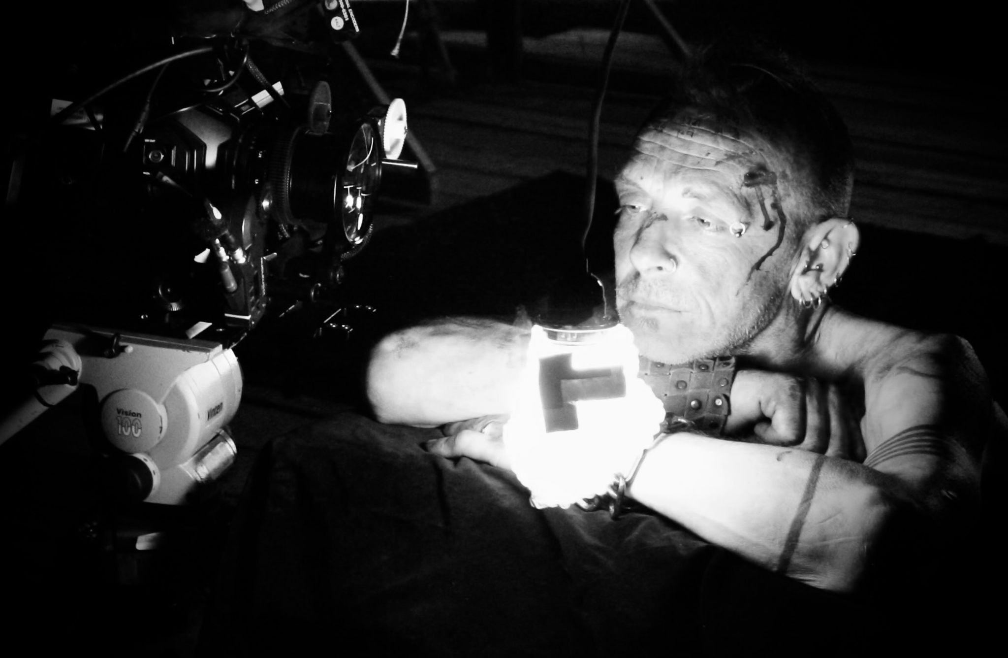 Black and white photo of a tattooed and pierced individual leaning forward with arms crossed, holding a bright light source that illuminates their face. A camera on a tripod is positioned in front of them, aimed at the subject. The background is dark, drawing focus to the person and the camera setup