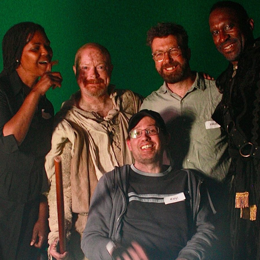 A group of five people pose in front of a green screen. One person, seated in a wheelchair at the center, wears glasses and a name tag. The other four stand around them, two on each side, dressed in various costumes, suggesting involvement in a production or performance. Focused lighting creates a bright spot at the bottom centre of the image