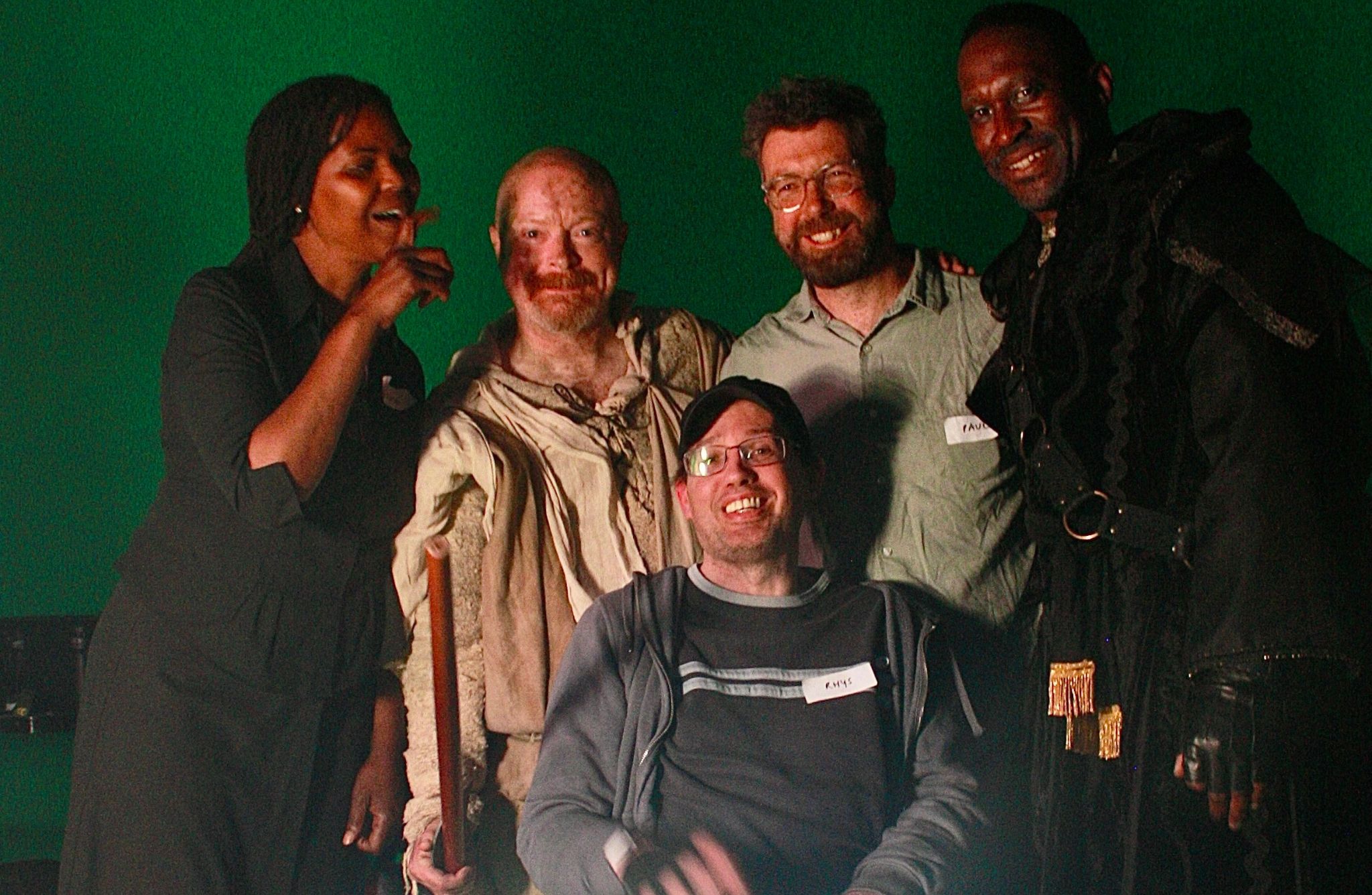 A group of five people pose in front of a green screen. One person, seated in a wheelchair at the center, wears glasses and a name tag. The other four stand around them, two on each side, dressed in various costumes, suggesting involvement in a production or performance. Focused lighting creates a bright spot at the bottom centre of the image
