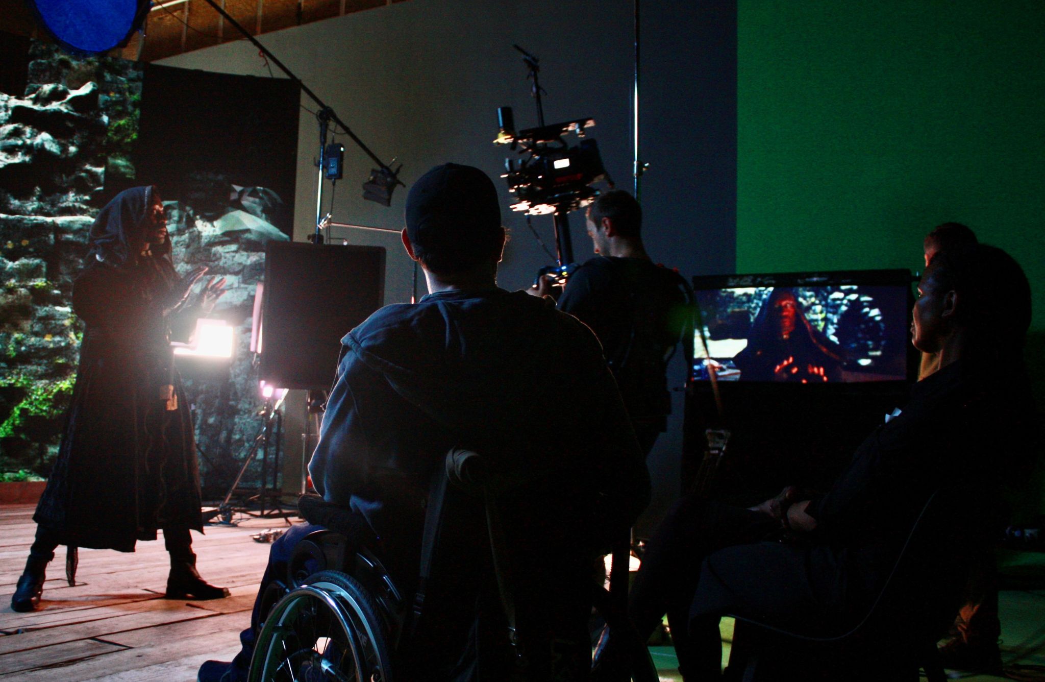 Behind-the-scenes view of a film set with four people. One person in a wheelchair faces away from the camera, while three others stand nearby, including one holding a camera rig. The set resembles a cave or rocky environment, with detailed rock formations in the background. A table in front holds props like skulls and other items suggesting an archaeological or adventure theme. Dramatic lighting enhances the atmosphere.