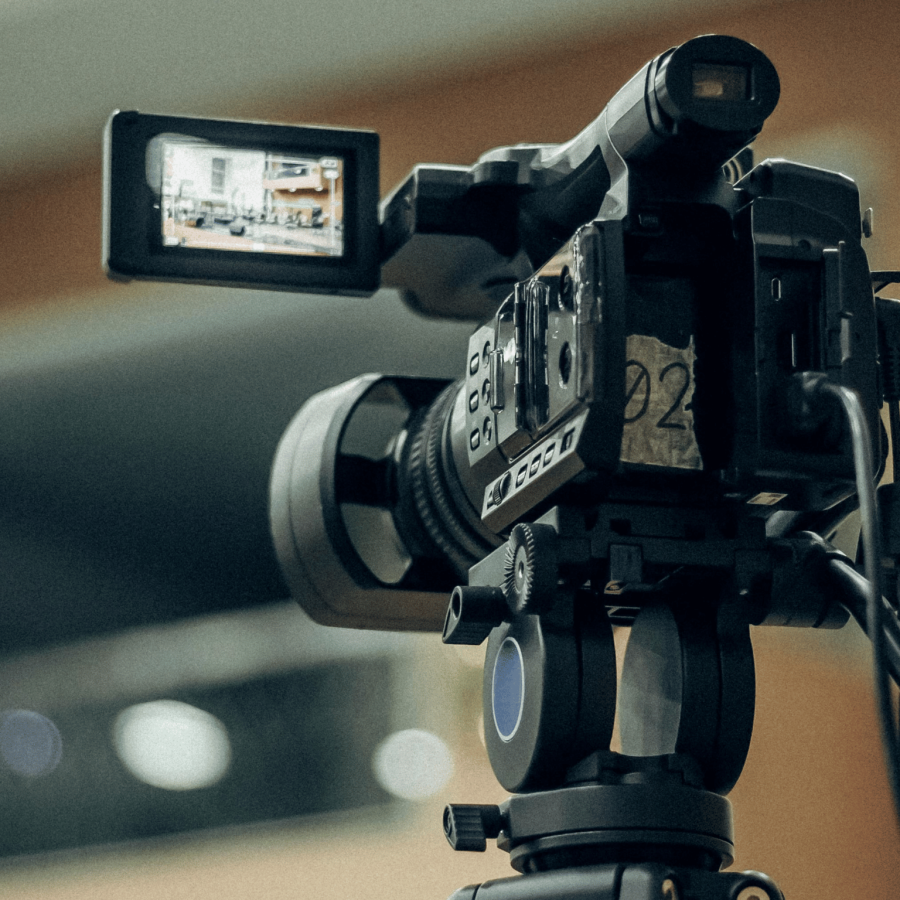 "Professional video camera mounted on a tripod, with an adjustable LCD screen displaying an indoor scene. The camera is equipped with control buttons, dials, and connected cables, emphasising its setup for recording."