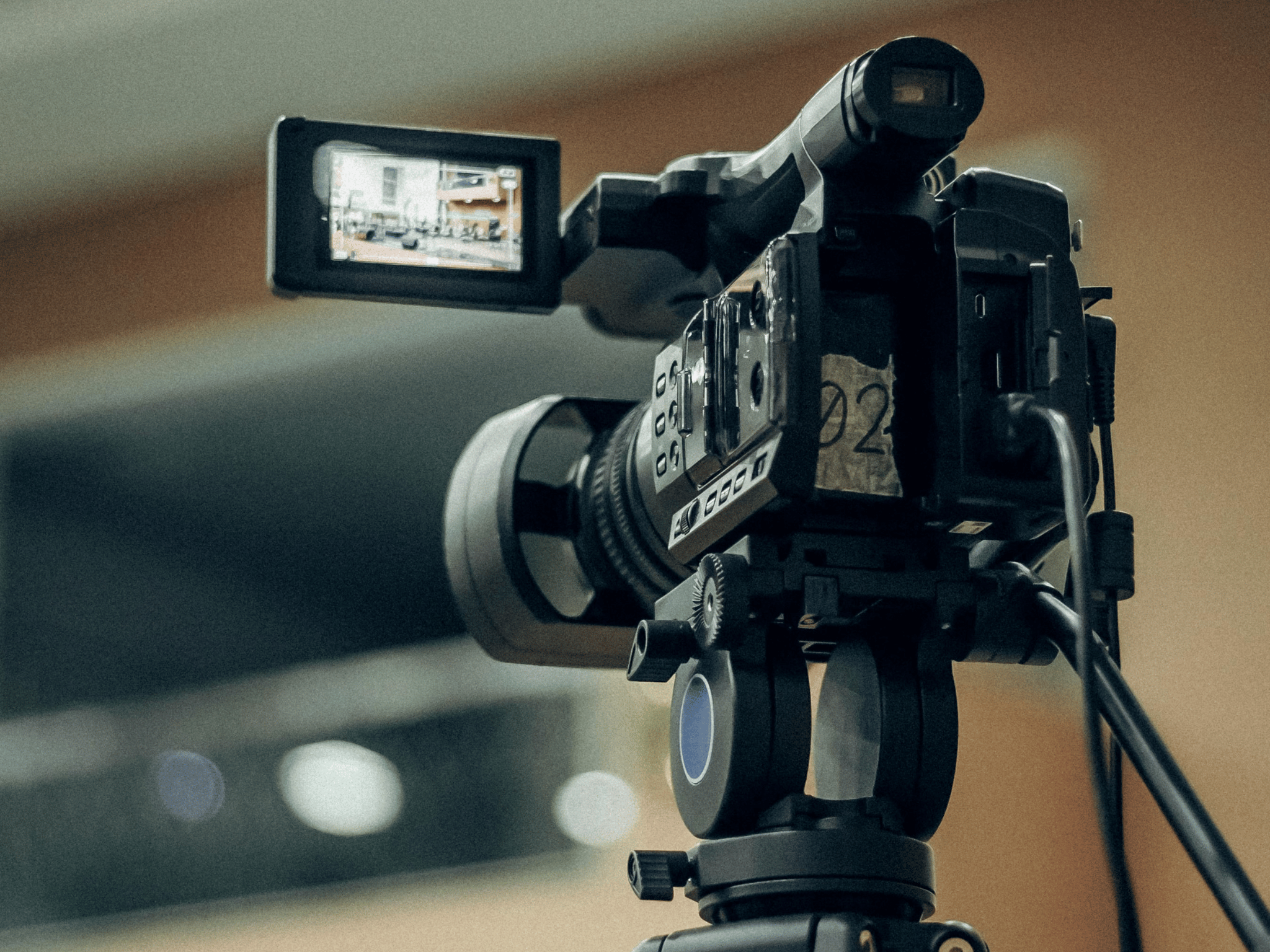 "Professional video camera mounted on a tripod, with an adjustable LCD screen displaying an indoor scene. The camera is equipped with control buttons, dials, and connected cables, emphasising its setup for recording."