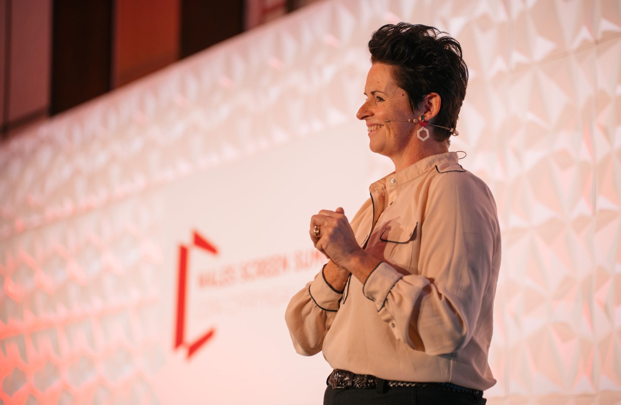 A person standing on a stage in front of a textured white backdrop with a red geometric logo and partially visible text that reads “WALES SCREEN” and “...”. The individual is wearing a light beige button-up shirt with rolled sleeves, dark trousers, and statement earrings, and appears to be speaking or presenting with hands clasped near the chest. Warm lighting highlights the scene